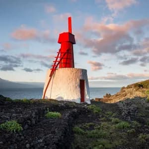 Un faro bianco e rosso su una costa vulcanica, con una montagna lontana e un mulino a vento rosso sullo sfondo.