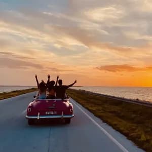 Un viaggio di gruppo WeRoad visto da dietro, alla guida di una decappottabile vintage rosa su una strada costiera durante un tramonto dorato.