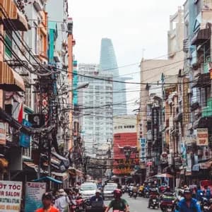 Eine belebte Stadtstraße voller Motorräder und Fußgänger, gesäumt von Gebäuden, einem Gewirr von Oberleitungen und einem modernen Wolkenkratzer im Hintergrund.