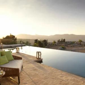 Un patio moderno con piscina a sfioro e mobili lounge che si affaccia su un paesaggio montano roccioso al tramonto.