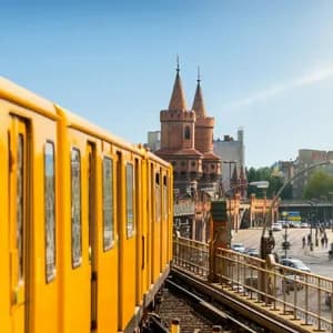 Un treno giallo viaggia su un binario sopraelevato attraverso un ponte, con un ponte storico della città e edifici sullo sfondo.