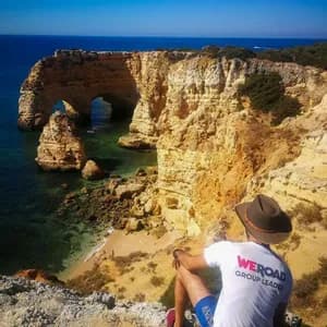 Un capo gruppo WeRoad con un cappello siede su una scogliera che si affaccia su una costa rocciosa con archi naturali sul mare e una piccola spiaggia.