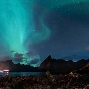 Das grüne Nordlicht leuchtet an einem sternklaren Nachthimmel über einem Fjord, mit Bergsilhouetten und einer felsigen Küste darunter.
