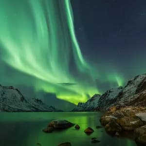 Corrientes verdes de la Aurora Boreal iluminan un cielo nocturno estrellado sobre un fiordo y montañas cubiertas de nieve.