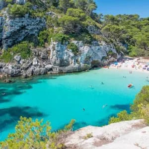 Vue aérienne d'une crique où des personnes nagent dans une eau turquoise claire et se détendent sur une plage de sable blanc entourée de falaises rocheuses et d'arbres.