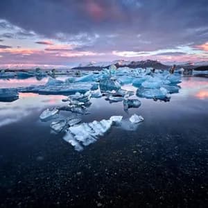 Iceberg galleggiano su acque calme riflettendo un cielo al tramonto, con montagne innevate in lontananza.