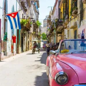 A pink vintage car parked on a narrow, sunlit street lined with colorful buildings, with a Cuban flag hanging from a balcony.