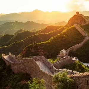 La Grande Muraille de Chine serpente à travers des montagnes verdoyantes et ondulantes, avec une tour de guet visible dans la lumière dorée d'un coucher de soleil.
