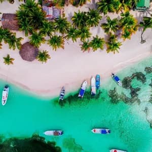 An aerial view of a WeRoad group trip on a white sand beach lined with palm trees, next to boats in turquoise water.