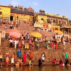 Una grande folla di persone in abiti colorati si raduna sui ghat di pietra, con molti che si bagnano nel fiume sotto tradizionali edifici gialli.