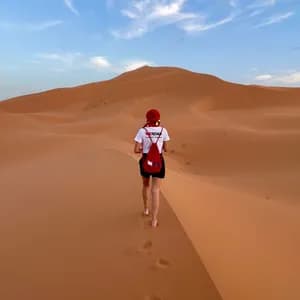 A person seen from behind wearing a WeRoad t-shirt and red cap walks barefoot up a large orange sand dune.