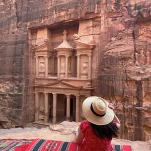 Una donna in abito rosso e cappello di paglia è seduta su un tappeto, osservando un grande tempio scavato in una scogliera di roccia rossa.