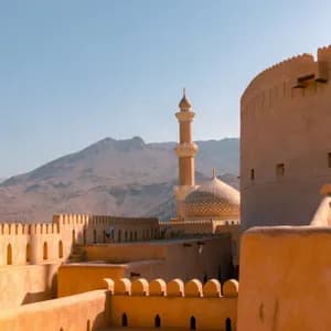A mosque's golden dome and minaret rise above the sand-colored walls of a fortress, with distant mountains under a clear sky.