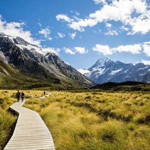Un voyage de groupe WeRoad se promène sur une passerelle en bois, traversant une vallée verdoyante, avec des montagnes enneigées en fond.
