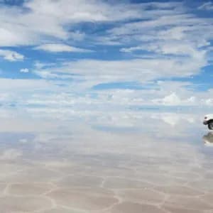 Un SUV bianco di un viaggio di gruppo WeRoad che attraversa una vasta e umida distesa di sale che riflette il cielo blu nuvoloso come uno specchio.