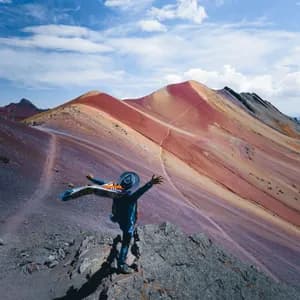 A person in a hat stands on a rocky outcrop with arms outstretched, overlooking a range of colorful, striped mountains under a blue sky.