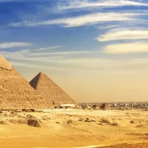 Vue panoramique de trois grandes pyramides dans un désert de sable avec une ville tentaculaire au loin sous un ciel bleu et nuageux.