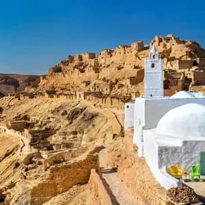 Un edificio imbiancato a calce con un minareto si erge in un villaggio desertico, incastonato in una collina rocciosa e assolata, sotto un cielo azzurro e limpido.