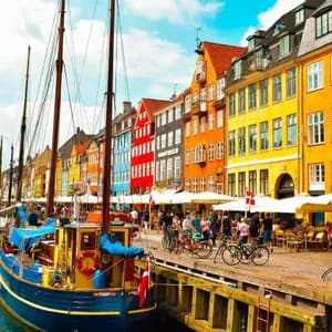 Bunte Gebäude säumen einen Kanal, an dem Segelboote festgemacht sind, während Menschen entlang der Uferpromenade spazieren und in Cafés sitzen.