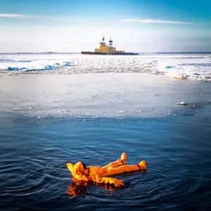Une personne en combinaison de survie orange flotte sur le dos dans l'eau glacée, avec un brise-glace au loin.