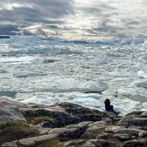 Una persona con i capelli rossi siede su una costa rocciosa, guardando un mare pieno di iceberg e ghiacciai sotto un cielo nuvoloso.
