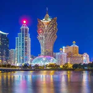 Grattacieli illuminati costeggiano un lungomare di notte, con un ponte sulla sinistra e luci colorate che si riflettono sull'acqua.