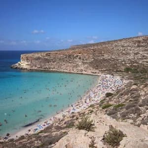 Una vista dall'alto di una spiaggia affollata in una cala rocciosa, con persone che nuotano in acque limpide color turchese sotto un cielo blu.