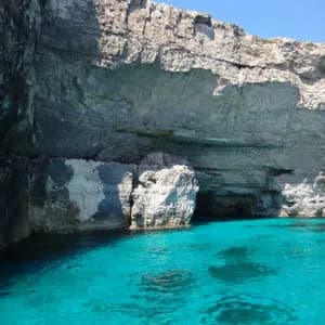 Una scogliera rocciosa con grotte marine alla base incontra acque turchesi brillanti sotto un cielo azzurro limpido.