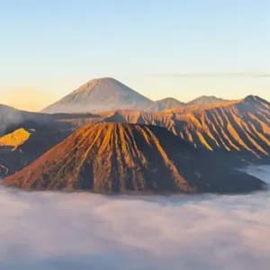 Un volcan fumant et d'autres sommets montagneux s'élèvent au-dessus d'une dense couche de nuages, illuminés par la lumière dorée du lever du soleil.