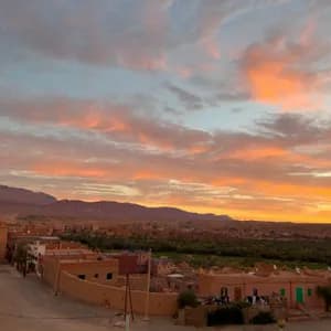 Un villaggio di edifici in terra si trova in una valle ai piedi delle montagne, sotto un cielo con nuvole arancioni e rosa al tramonto.