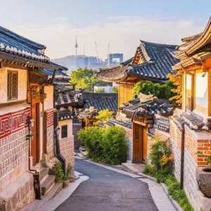 Una stretta via si snoda tra case tradizionali con tetti in tegole, con lo skyline moderno di una città e una torre visibili sullo sfondo.