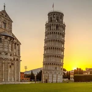 Ein schiefer Glockenturm und eine kunstvolle Kathedrale stehen auf einem grünen Rasen vor einem goldenen Sonnenuntergang.