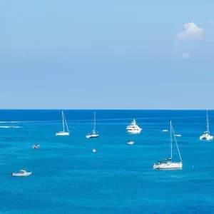 Vista panoramica di diverse barche a vela su un mare blu intenso, accanto a una grande scogliera rocciosa, sotto un cielo limpido.