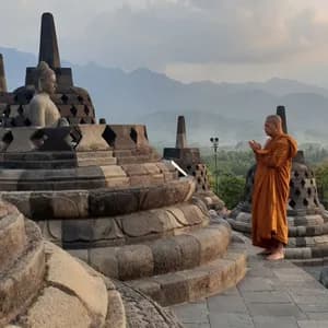 A Buddhist monk in orange robes prays among ancient stone stupas with a mountain landscape in the background.