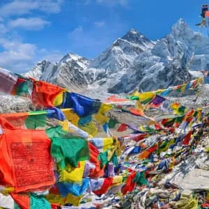 Colorful prayer flags stretched across a rocky viewpoint, with snow-capped mountains and a glacier in the background under a blue sky.