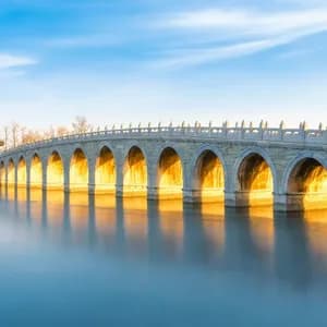 Un lungo ponte di pietra ad arco illuminato dalla luce dorata del sole attraversa acque calme, con il suo riflesso visibile sulla superficie.