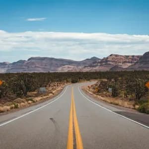 Una strada a due corsie serpeggiante che attraversa un paesaggio desertico con alberi di Joshua e montagne rocciose sotto un cielo parzialmente nuvoloso.