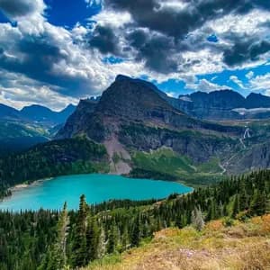 Un lago turchese brillante riposa in una valle, circondato da una fitta foresta sempreverde e aspre montagne sotto un cielo azzurro nuvoloso.