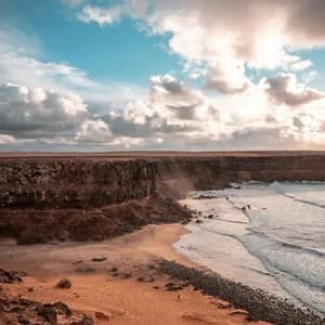 Ein Blick von oben auf eine felsige Küste mit orangem Sand, dunklen Klippen und Meereswellen unter einem teilweise bewölkten Himmel.