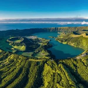 Veduta aerea di una grande caldera vulcanica contenente un lago azzurro, circondata da rigogliose colline verdi, con l'oceano visibile all'orizzonte.
