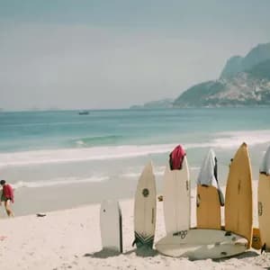 Auf einem sonnigen Strand stehen mehrere Surfbretter aufrecht im Sand. Im Hintergrund entspannen Menschen am Meer vor einer Bergkulisse.