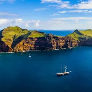 A panoramic aerial view of a rugged, green coastline and deep blue sea, with a large sailing ship and other boats.