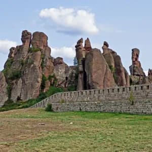 Una fortezza di pietra costruita in imponenti formazioni rocciose rossastre su una collina erbosa sotto un cielo parzialmente nuvoloso.
