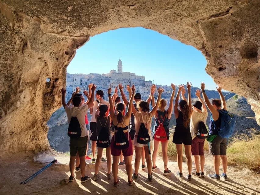 Un gruppo WeRoad, ripreso di spalle, alza le mani all'interno di una grotta con vista su una città storica in collina.
