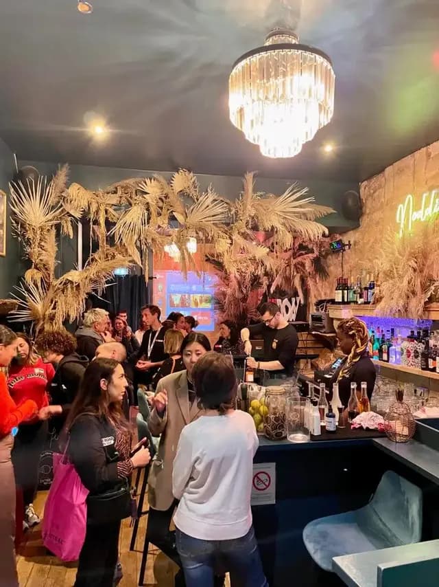 Un folto gruppo di persone che socializza in un bar affollato ed elegante, con un barista che prepara bevande sotto un grande lampadario.