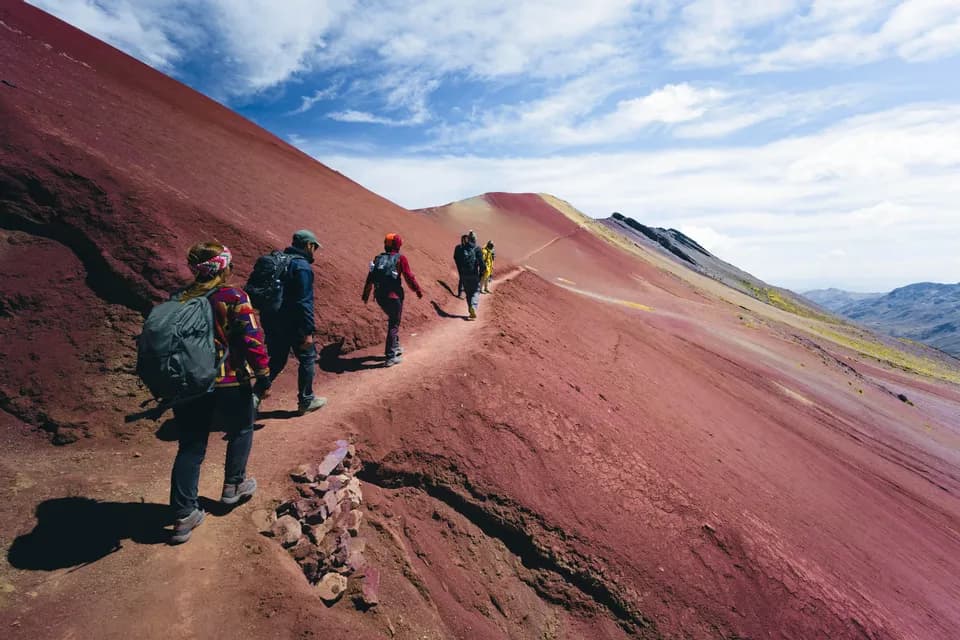 Un viaje en grupo de WeRoad camina en fila india por un sendero estrecho en una montaña roja y empinada bajo un cielo azul.