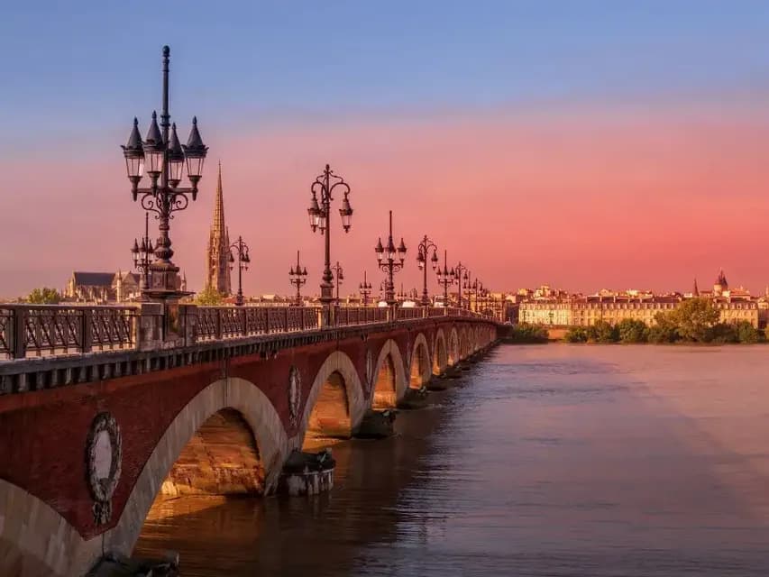Un ponte ad arco in mattoni con lampioni ornamentali attraversa un fiume al tramonto, con uno skyline cittadino sullo sfondo.