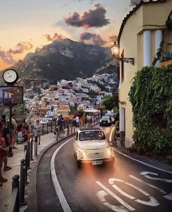 Un coche clásico con las luces encendidas sube por una carretera sinuosa a través de un pueblo en el acantilado, con montañas al fondo al atardecer.