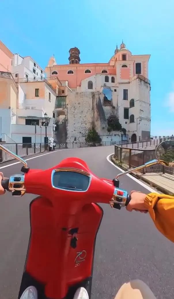 Vista en primera persona de un paseo en scooter rojo por una carretera sinuosa hacia un pueblo costero con edificios históricos.