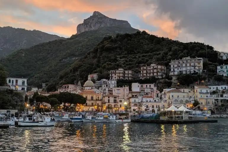 Un pueblo costero con un puerto lleno de barcos anidado al pie de una montaña verde al atardecer.